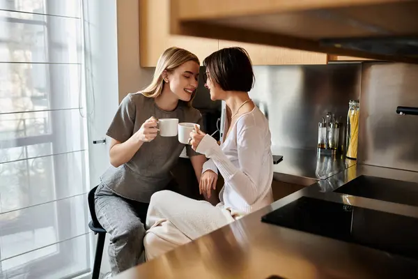 Молода лесбійська пара пов'язана з чашками кави, сидячи поруч і спілкуючись у затишній кухні всередині готелю. — стокове фото