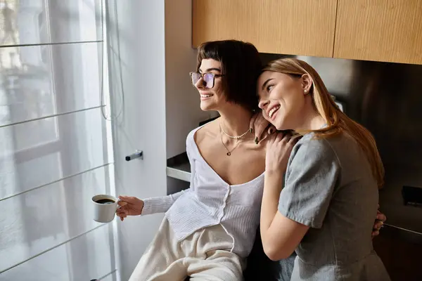 Two young women, a lesbian couple, standing close with coffee inside a hotel room kitchen. — Stock Photo