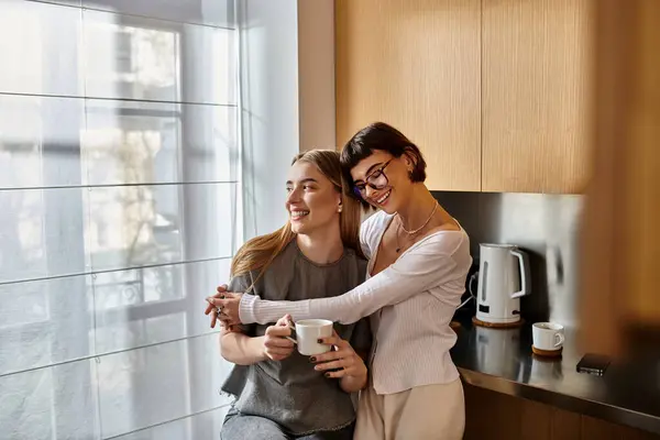 Dos mujeres jóvenes, una elegante pareja de lesbianas, se paran una al lado de la otra en una moderna cocina del hotel, sosteniendo tazas de café. - foto de stock