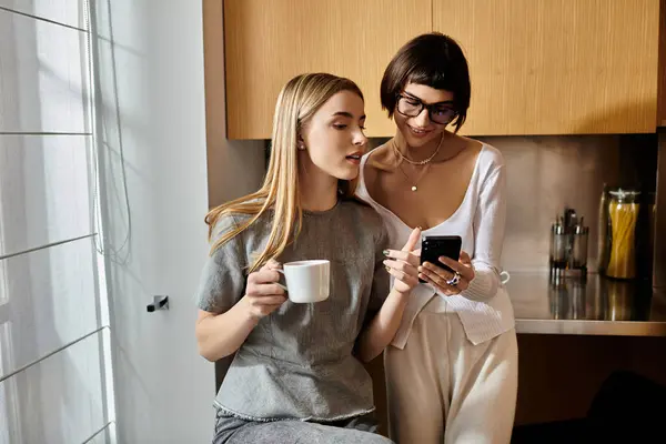 A young lesbian couple sit on a kitchen counter, each engrossed in smartphone, holding coffee cup. — Stock Photo