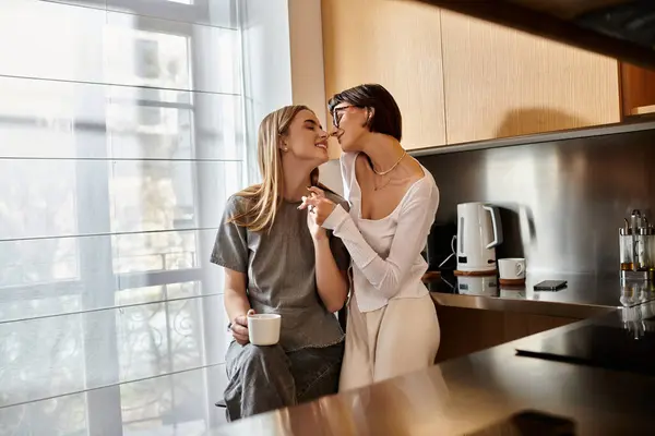 Two young women, a lesbian couple, stand side by side in a kitchen with coffee cups in hand, starting their day. — Stock Photo