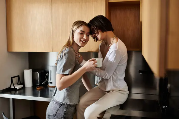 Un hombre y una mujer jóvenes, una pareja lesbiana, sentados en un mostrador de cocina en una habitación de hotel, tomando café. - foto de stock