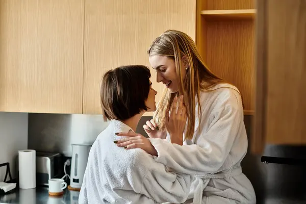 Two women in bath robes standing together, sharing an intimate moment in a warm and inviting kitchen setting. — Stock Photo