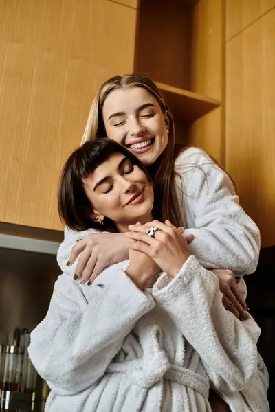 Due giovani donne, una coppia lesbica, avvolte in accappatoi, condividono un caldo abbraccio in una cucina accogliente. — Foto stock