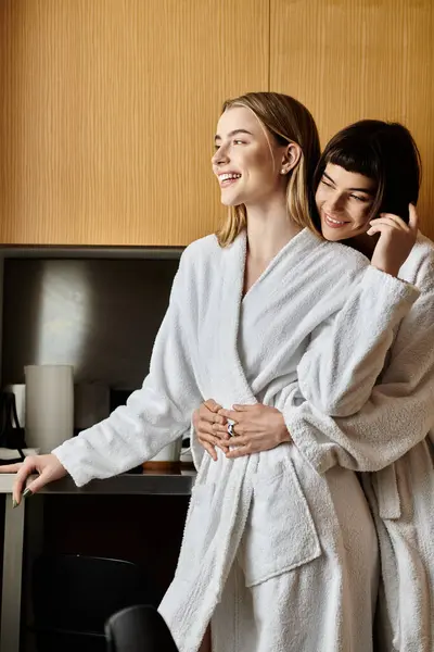 Duas jovens mulheres em roupões de banho de pé perto, exalando amor e conexão em um ambiente acolhedor quarto de hotel. — Fotografia de Stock