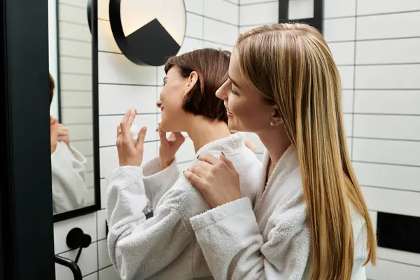 Две молодые женщины в белых халатах стоят перед зеркалом в ванной комнате отеля, разделяя нежный момент. — стоковое фото