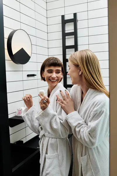 Zwei junge Frauen in Bademänteln stehen vor einem Badezimmerspiegel und starren einander mit Zuneigung an. — Stockfoto
