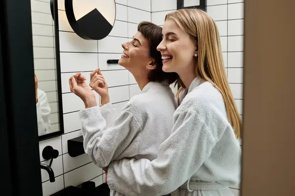 A couple of women in bath robes stand together, sharing a tender moment in front of a bathroom mirror. — Stock Photo