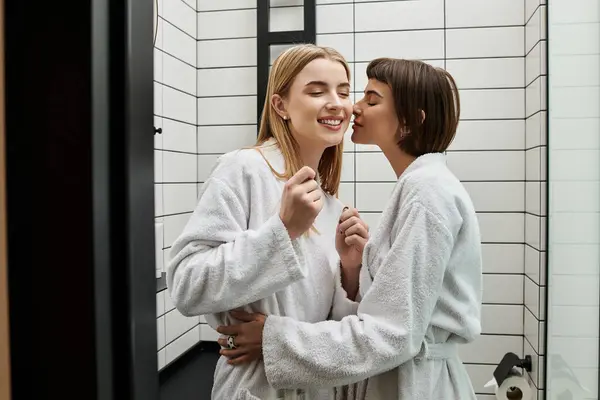 Una giovane coppia lesbica in accappatoi, in piedi in bagno, baci guancia — Foto stock