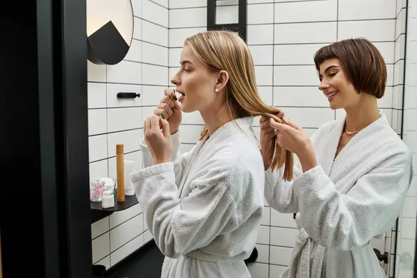 A young lesbian couple in bath robes, one flossing teeth, other braiding hair in a hotel bathroom. — Stock Photo