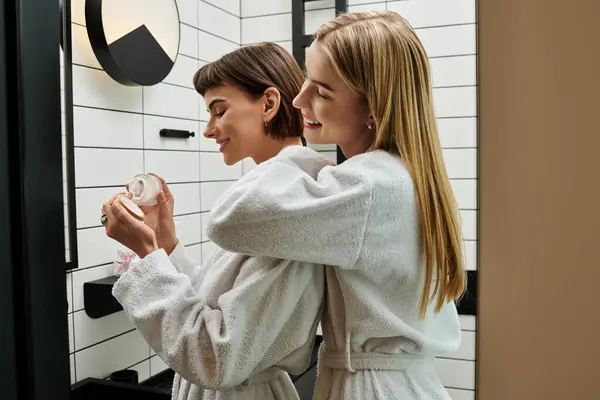 Duas jovens mulheres em vestes brancas olham para o seu reflexo em um espelho em um banheiro do hotel. — Fotografia de Stock