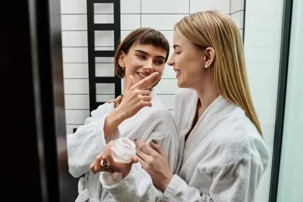 Dos mujeres jóvenes con batas de baño aplicando crema en un baño de hotel. - foto de stock