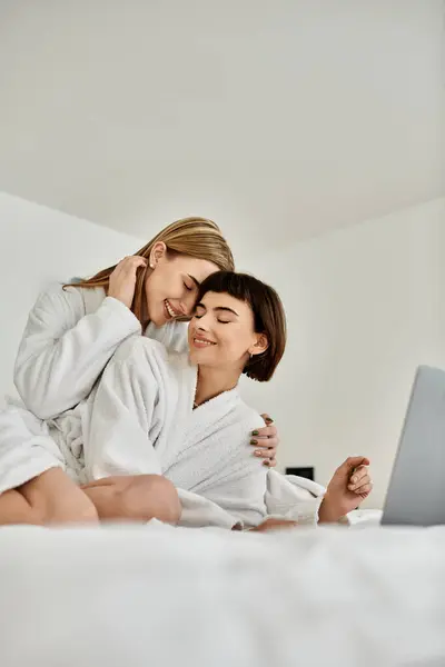 A young lesbian couple in bath robes relaxes on top of a cozy bed in a hotel room. — Stock Photo