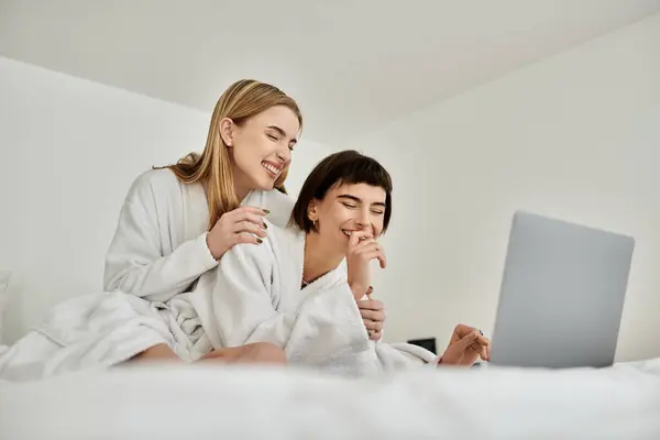 Duas jovens mulheres em roupões de banho sentam-se confortavelmente em uma cama luxuosa, compartilhando um momento terno juntas. — Fotografia de Stock