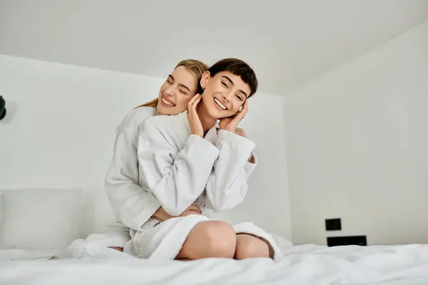 A beautiful lesbian couple in bath robes sit peacefully on top of a cozy bed inside a hotel room. — Stock Photo