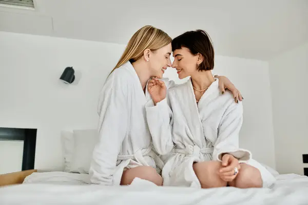 A beautiful lesbian couple in white robes sit serenely on a bed inside a hotel room, exuding grace and peace. — Stock Photo