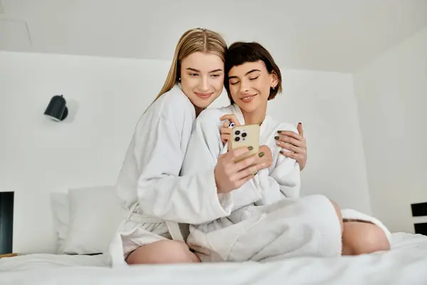 Две женщины в халатах сидят на кровати, очарованные экраном мобильного телефона. — стоковое фото