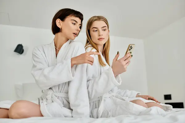 Two beautiful women, in bath robes, relax and chat on a hotel bed. — Stock Photo