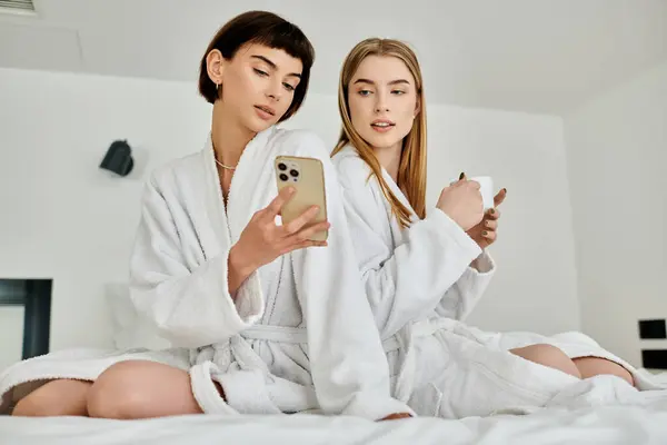 A beautiful lesbian couple dressed in bath robes sitting on top of a bed in a hotel room, enjoying a moment of relaxation. — Stock Photo