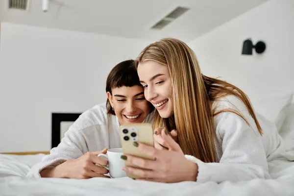 A beautiful lesbian couple in bath robes lay in bed, deeply engrossed in the soft glow of a cell phone screen. — Stock Photo