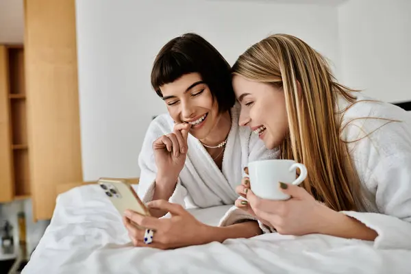 Two beautiful lesbian women in bath robes lying on bed, engrossed in content on a smartphone. — Stock Photo