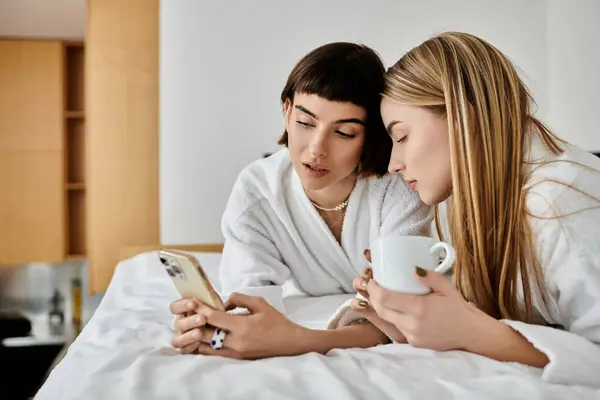 A beautiful lesbian couple in bath robes lay on bed, engrossed in cell phone — Stock Photo