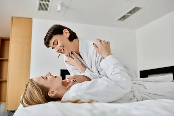 Um belo casal de lésbicas de roupões de banho deitados um ao lado do outro em uma cama dentro de um quarto de hotel. — Fotografia de Stock