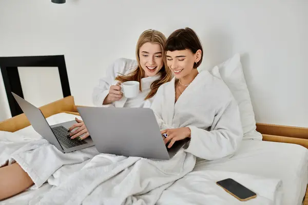 Two women in white robes collaborate on a laptop while lounging on a comfortable bed inside a hotel room. — Stock Photo