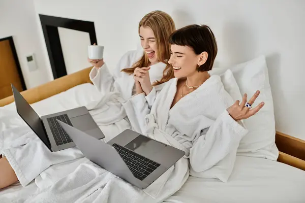 Two beautiful women, a lesbian couple, sit on a bed, each with a laptops, deep in conversation and digital communication. — Stock Photo