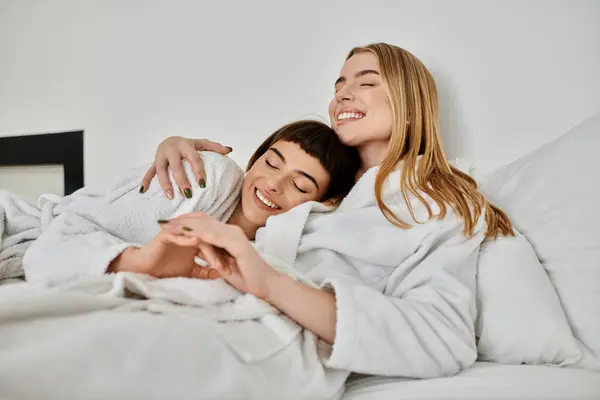 A beautiful lesbian couple in bath robes peacefully laying on top of a comfortable bed inside a hotel room. — Stock Photo
