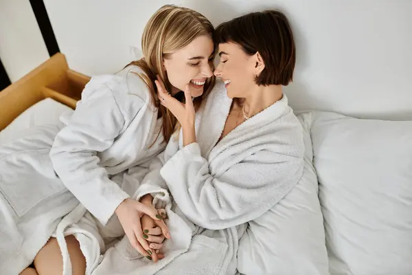 A relaxed lesbian couple in bath robes sit atop a neatly made hotel bed, enjoying a quiet moment together. — Stock Photo
