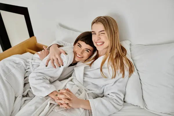 A beautiful lesbian couple in bath robes lying together on a plush hotel bed, sharing a tender moment. — Stock Photo