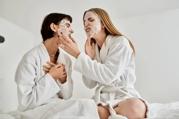 A beautiful lesbian couple in white robes sitting peacefully on a bed inside a hotel room. — Stock Photo