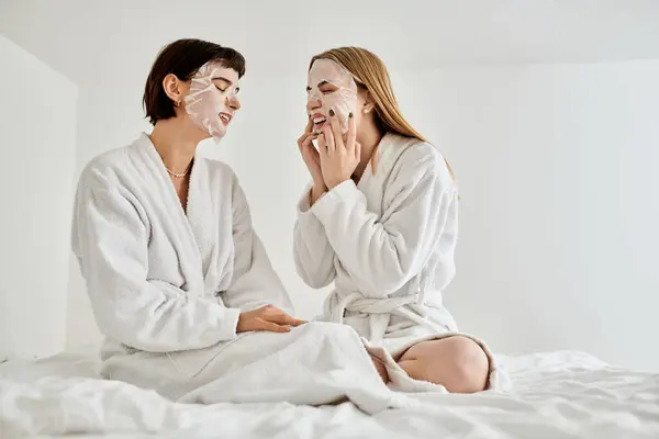 A beautiful lesbian couple in sheet masks and bath robes peacefully sitting on a luxurious hotel bed together. — Stock Photo