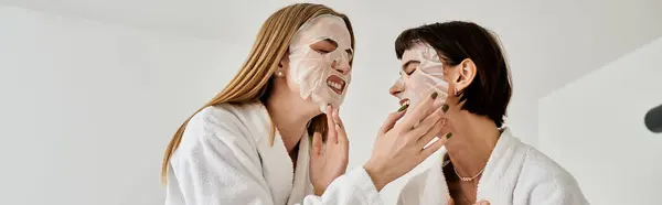 Two beautiful women in bath robes relaxing with facial masks on their faces in a cozy hotel room. — Stock Photo