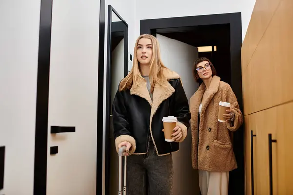 Um jovem casal lésbico com bagagem, elegantemente de pé juntos em um corredor do hotel. — Fotografia de Stock