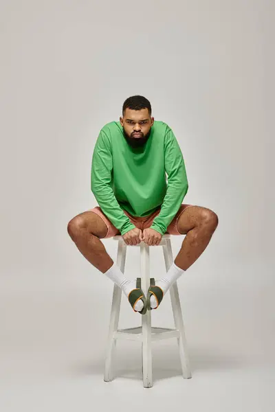 Handsome African American man sitting on stool in stylish green shirt against vibrant backdrop. — Stock Photo