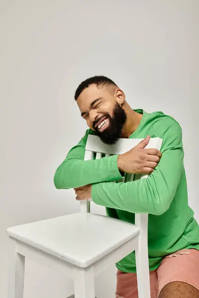 A stylish African American man sits confidently on a white chair against a vibrant backdrop. — Stock Photo