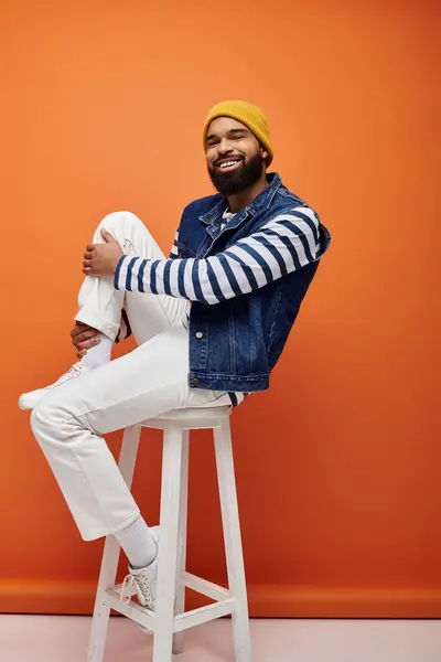 Handsome African American male in trendy outfit sitting atop white stool against colorful background. — Stock Photo