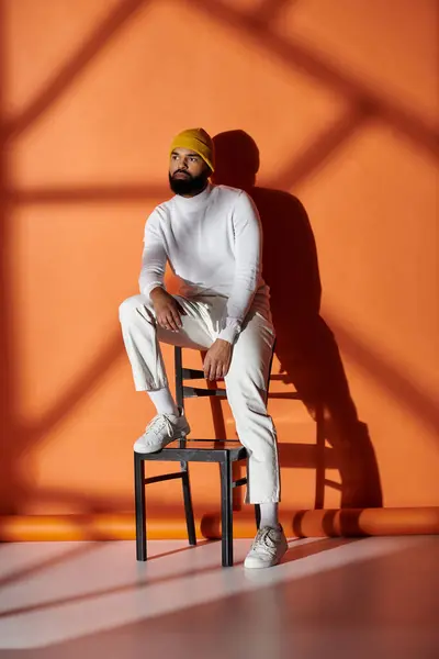 Handsome African American man in fashionable attire sitting on top of a chair in front of a vibrant orange wall. — Stock Photo