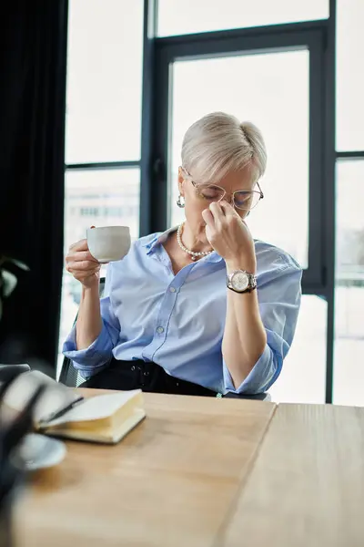 Middle-aged businesswoman with short hair enjoying a cup of coffee at a table in an office setting. — Stock Photo