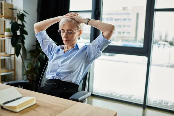 Une femme d'affaires d'âge moyen est assise au bureau, les mains sur la tête, profondément dans la pensée. Cheveux courts et tenue professionnelle. — Photo de stock