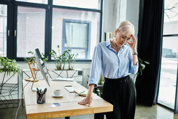 Una donna d'affari di mezza età con una camicia blu si trova a una scrivania in un ufficio, sentendosi male durante la menopausa. — Foto stock