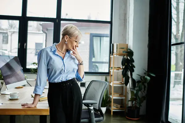 Middle-aged businesswoman in blue shirt and black pants stands at desk, feeling unwell during menopause. — Stock Photo