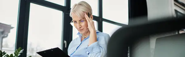 Empresária de meia-idade com cabelo curto visivelmente lutando, sentada na mesa com a mão na cabeça durante a menopausa no escritório. — Fotografia de Stock
