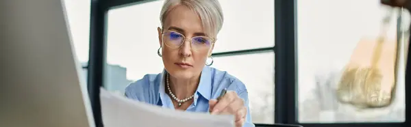 A focused middle-aged businesswoman with short hair scrutinizing a piece of paper while seated in front of a computer. — Stock Photo
