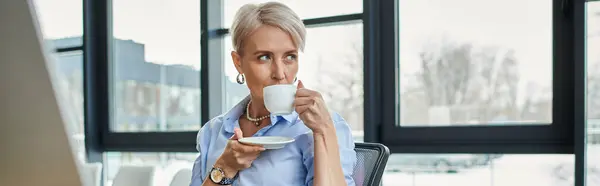 Empresária de meia-idade com cabelo curto desfruta de um momento tranquilo, sentada em sua mesa e tomando uma xícara de café. — Fotografia de Stock