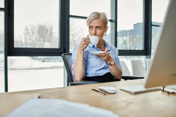 Donna d'affari di mezza età, capelli corti, che lavora durante la menopausa, si siede alla scrivania, si gode una tazza di caffè. — Foto stock