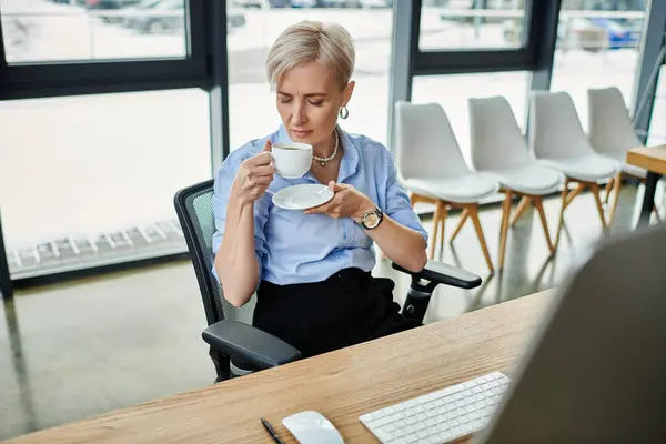 A middle-aged businesswoman with short hair sitting at a desk, enjoying a cup of coffee while working in an office. — Stock Photo