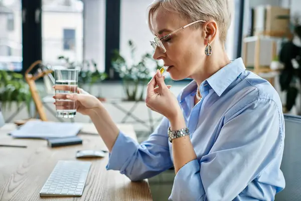 Middle-aged businesswoman with short hair working at desk, taking pill during menopause. — Stock Photo